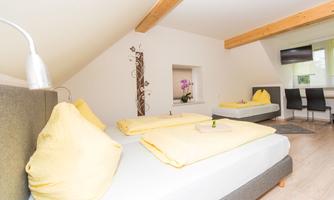 A bright bedroom with two beds and yellow pillows. There is a table and chairs as well as a television on the wall.