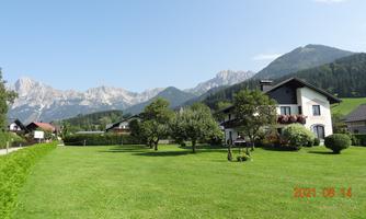A picturesque landscape with a large green garden and a white house. In the background, majestic mountains and a clear sky can be seen.