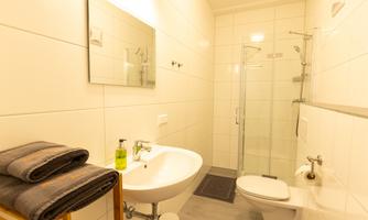 A modern bathroom with a shower, a sink, and a toilet. The walls are tiled in light colors, and towels are neatly placed on a shelf.