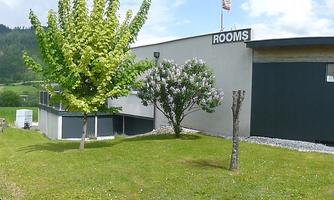 A modern building with the inscription "ROOMS" stands in a green landscape. Gentle hills and a blue sky are visible in the background.