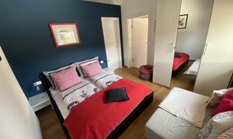 A cozy bedroom with a double bed and colorful pillows. In the background, there is a small seating area and a wardrobe.