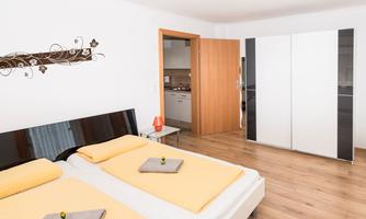 A modern bedroom with two single beds and yellow bed sheets. In the background, an open kitchen corner can be seen.