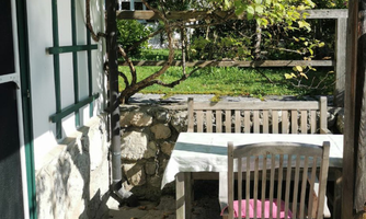 An inviting outdoor area with a wooden table and chairs, surrounded by nature. The sun illuminates the space and creates a cozy atmosphere.