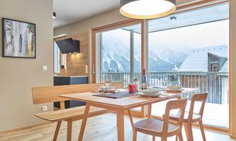 A modern dining room with a wooden chair and a table set for a meal. The large windows offer a view of the mountains.
