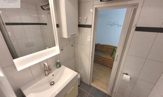 A modern bathroom with white tiles and a large mirror. There is a sink, a door to another room, and a window for natural light.