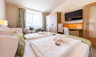 A modern hotel room with two beds and a large window. The room is bright and tastefully furnished, with a seating area and a television.