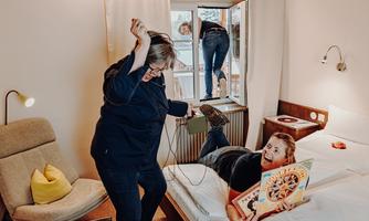 A humorous situation in a hotel room, where one person is dancing with an electric device while another is lying on the bed reading a book. In the background, someone is trying to climb out of the window.