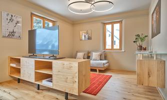 A modern living room with a TV on a wooden cabinet. There is a cozy sofa and large windows that let in plenty of light.