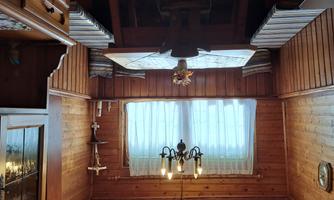 A cozy room with wood-paneled walls and a large table. There are windows with curtains and a ceiling lamp that creates an inviting atmosphere.