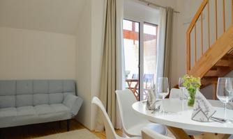 A bright and modern living room with a gray sofa and a round dining table. Large windows provide plenty of daylight.