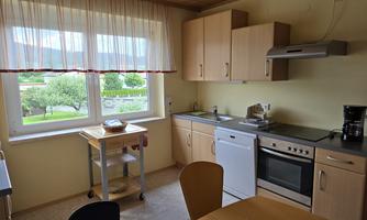 A bright kitchen with wooden cabinets and a table. A large window offers a view of the green landscape outside.