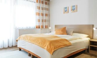 A cozy bedroom with a large bed and an orange throw. The walls are kept light, and there are windows with curtains as well as two pictures on the wall.