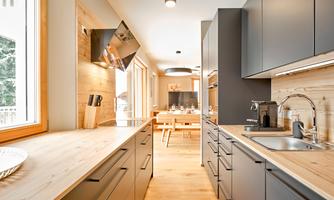 A modern kitchen with wooden shelves and elegant black kitchen furniture. Plenty of natural light streams through the windows, creating an inviting atmosphere.