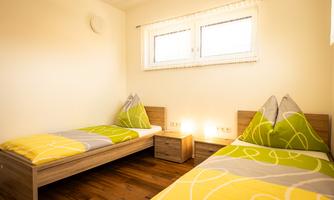 A bright bedroom with two single beds and colorful bedcovers. The windows provide plenty of daylight, and there is a small bedside table between the beds.