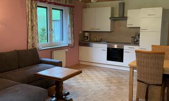 A cozy living room with a gray sofa, a wooden coffee table, and a dining table. The kitchen is modern and well-equipped, featuring white cabinets and various appliances.