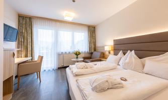A modern hotel room with two beds, a television, and a seating area. Large windows provide plenty of light and a view outside.