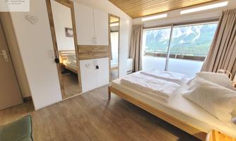A modern bedroom with a large window and a view of the mountains. There is a double bed, a wardrobe, and cozy furnishings.