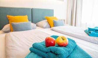 A cozy bedroom with two beds, colorful cushions, and fresh apples on a folded towel. The room has a bright and inviting atmosphere.
