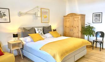 A cozy bedroom with a large bed and yellow cushions. A wooden wardrobe and a plant add warmth and style to the room.