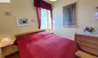 A cozy bedroom with red bedding and a light wall. The room is well-lit and decorated with pictures and a floral arrangement.