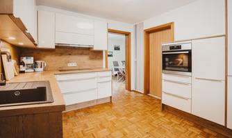 A modern kitchen with white cabinets and wood details. The floor is covered with parquet and there is plenty of space for activities.