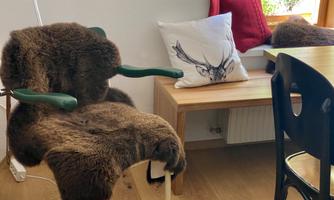 A cozy chair with a fur cover is next to a wooden table. On the table lies a cushion with a stylized deer motif.