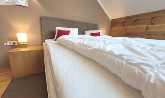 A modern bedroom with a large bed and wooden elements. The bedding is white, and there is a cozy bedside lamp.