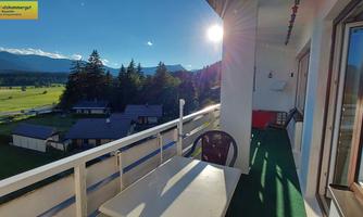 A balcony with a table and a chair, overlooking a green landscape and the mountains in the background. The sun is shining brightly, creating a pleasant atmosphere.
