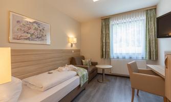 A simple and modern room with a single bed and a desk. Large windows provide plenty of daylight and a pleasant atmosphere.
