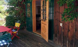 A wooden terrace with colorful chairs and an open entrance to a cozy room. Surrounded by plants and natural beauty.