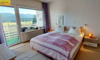 A cozy bedroom with a large bed and bright doors that lead to a balcony. The windows offer a view of the mountains and the landscape outside.
