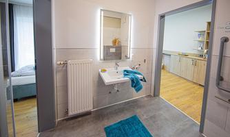 A modern bathroom with a sink, a large mirror, and a towel in bright blue. The room has light walls and a wooden floor, while an adjacent room is visible.