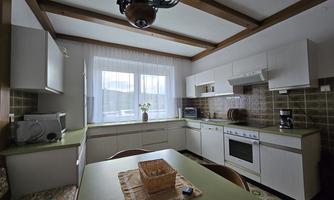 A bright kitchen with a green table and white furniture. There is a window with curtains and some kitchen appliances in the background.