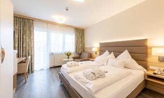 A modern hotel room with a large double bed and two pillows. The room is bright and inviting, with large windows and a cozy seating area.