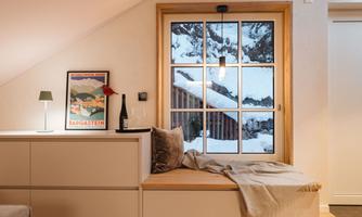 A cozy room with a window through which you can see snow-covered mountains. On the windowsill lies a blanket, and there are a few decorative items.