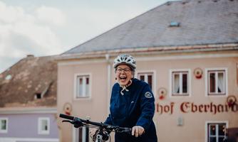 A cheerful woman on a bicycle is smiling. In the background, a historic building can be seen.