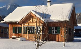 A charming wooden house in the snow, surrounded by snow-covered trees. The sun shines on the detailed facade decoration and the green shutters.