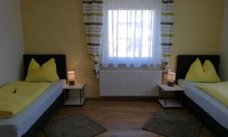 A cozy bedroom with two single beds and yellow pillows. Bright walls and a window provide plenty of daylight.