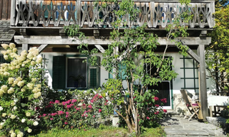 A cozy house with a balcony and blooming flowers in the foreground. The facade is bright and friendly, surrounded by plants.
