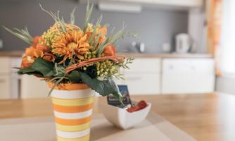 A colorful bouquet in a striped pot stands on a wooden table. In the background, a modern kitchen with white dishes and a kettle can be seen.