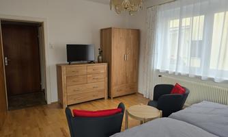 A bright bedroom with a double bed, a television, and a wooden wardrobe. Next to it are two armchairs and a small table.