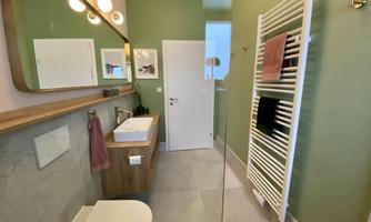 A modern bathroom with green walls and a large mirror. It features a shower, a wooden sink, and a towel radiator.