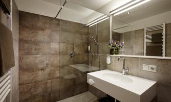 A modern bathroom with a glass shower and a simple, white sink. The walls are covered with brown tiles, and there are some plants for decoration.