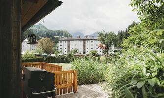 A cozy terrace with a grill and lots of green foliage. In the background, several residential buildings and mountainous landscapes are visible.