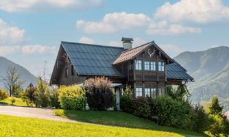 A charming wooden house surrounded by green meadows and trees. In the background, gentle hills and a blue sky can be seen.