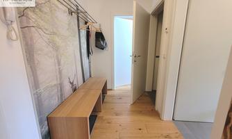 A bright hallway with wooden flooring and a bench. Clothing items hang on the wall, and an open room is visible.