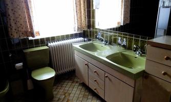 A bathroom with a double sink and a bathtub in shades of green. Bright windows and simple wooden furniture complement the space.