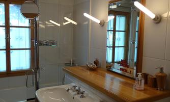 A modern bathroom with a shower and a wooden sink. The walls are light-tiled and there are large windows that let in lots of light.