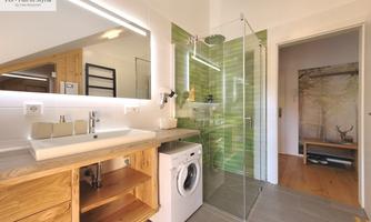 A modern bathroom with a glass enclosure and a washing machine. The space is bright and designed with wood and green tiles.