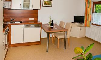 A modern kitchen with a small dining area and a table. Wine and glasses are on the table, and a television is located on a sideboard.